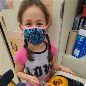 Kindergarner with mask on smiling at the camera as she digs out the inside of a pumpkin 